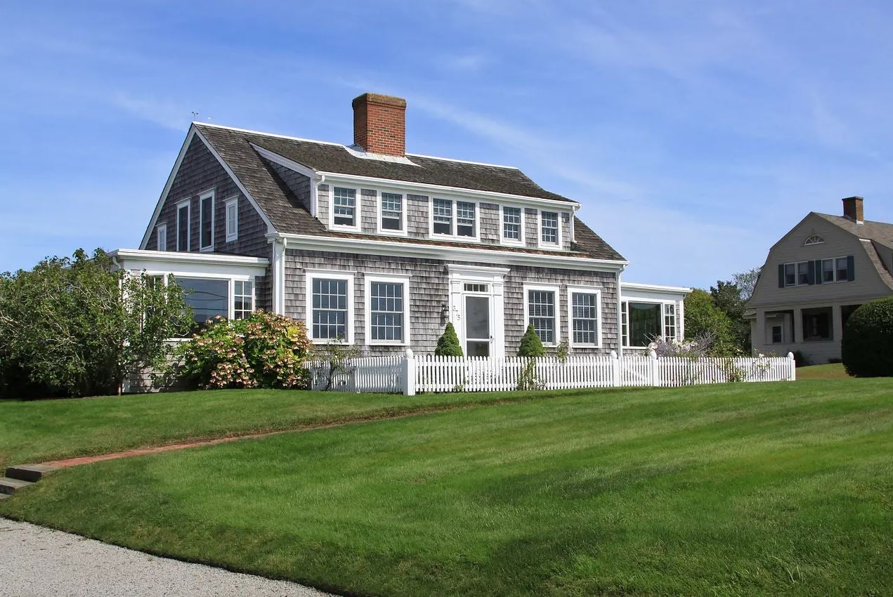 A Cape Cod style home with a large front lawn, wooden siding and a grey roof