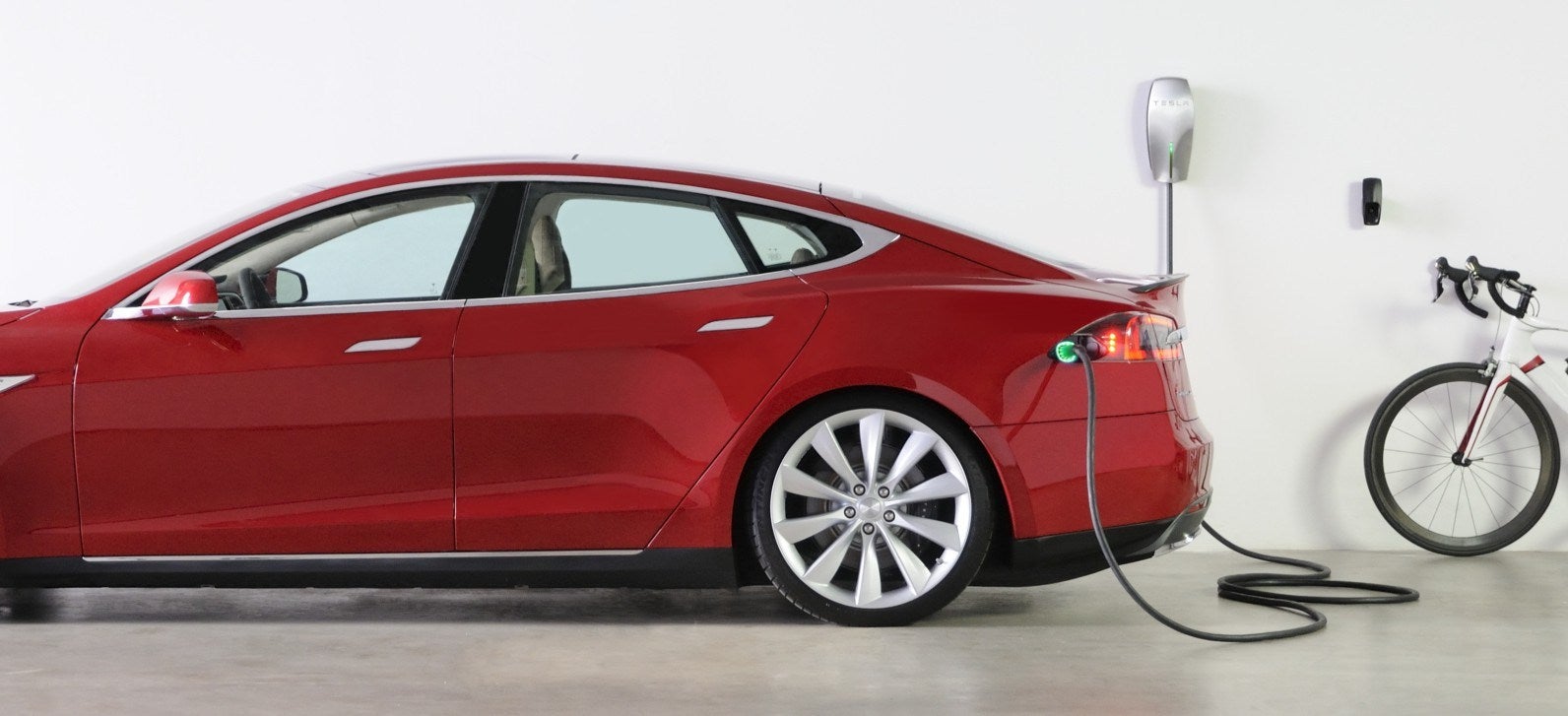 A Tesla Model S sedan charging from a wall connector in a garage.